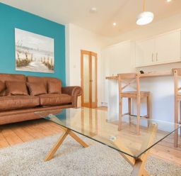 A living room with a brown sofa a glass coffee table and a kitchen counter with wooden stools at The Loft (Amble) in Amble