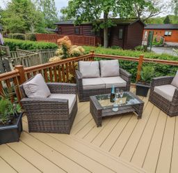 An outdoor wooden deck with rattan sofa and chairs around a glass coffee table with a bottle and glasses at 8 Waterside Wood White Cross Bay near Troutbeck Bridge