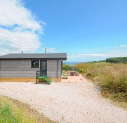 A small gray house with green steps and a tiled patio in a rural area with hills and blue sky at Midway Lodge in Cawsand near Kingsand