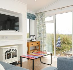 A living room with a television above a white electric heater two blue sofas and a colorful coffee table by a wooden side table with a framed picture and large windows with floral curtains at Midway Lodge in Cawsand near Kingsand