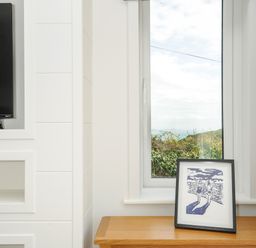 A window with a view of greenery and a body of water beside a wooden cabinet with a framed artwork a TV on a white wall in Midway Lodge in Cawsand near Kingsand