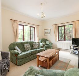 A living room with green sofas and armchair a wooden coffee table a TV on a stand and floral curtains at Brantfell Lodge in Bowness-On-Windermere