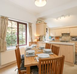 A dining area with wooden table and six chairs set with dishes beside a kitchen with light wood cabinets at Brantfell Lodge in Bowness-On-Windermere