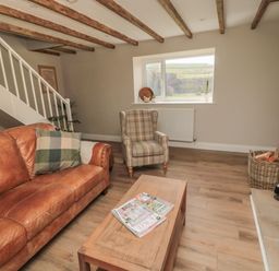 A living room with a leather sofa a plaid armchair a wooden coffee table magazines on the table a window stairs and a basket with firewood at The Hollow in West Woodburn