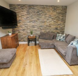 A living room with a gray sectional sofa, a wooden cabinet with a plant, a wall-mounted television, and a small table with a plant at Fir Tree Cottage in Llanrug