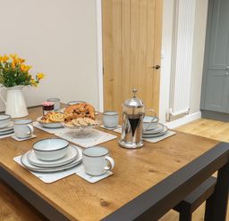 A dining table set with plates mugs a French press and baked goods in a kitchen at Fir Tree Cottage in Llanrug