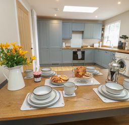 A kitchen with blue cabinets and a wooden dining table set with dishes pastries and a vase of yellow flowers at Fir Tree Cottage in Llanrug
