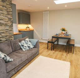 A living area with a grey sofa and patterned pillows a wooden dining table with benches and kitchen cabinets at Bay Tree Cottage in Llanrug