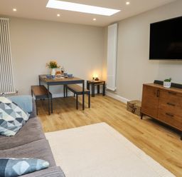 A living room with a sofa pillows dining table bench wooden cabinet and wall mounted tv at Bay Tree Cottage in Llanrug