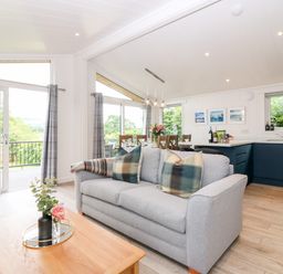 A living room with a gray sofa and plaid pillows a wooden coffee table with flowers a dining area with a wooden table and chairs and a kitchen with blue cabinets at Eden Fields Lodge in Cupar Fife