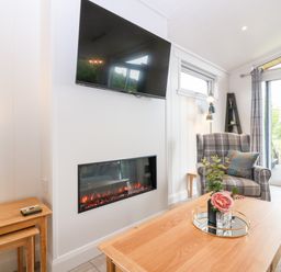 A living room with a wall-mounted television an electric fireplace a plaid armchair a wooden coffee table with flowers and a side table near a large window at Eden Fields Lodge in Cupar Fife