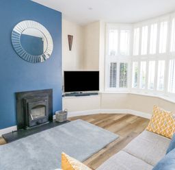 A living room with a blue accent wall fireplace a television on a corner shelf bay windows with white shutters and a gray sofa with yellow and blue cushions at Leos Place in Weymouth