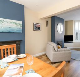 A dining and living area with a wooden table set for a meal a beige armchair a television and shuttered windows at Leos Place in Weymouth