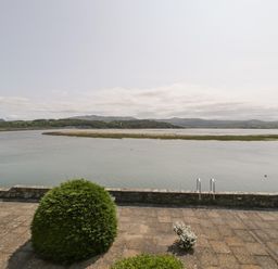 A waterfront view with a stone patio, bushes, a low stone wall, water, and hills in the background at 25 South Snowdon Wharf in Porthmadog