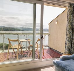 A wooden balcony with two folding chairs and a small table with a book overlooking a body of water seen through a sliding glass door next to a light blue armchair at 25 South Snowdon Wharf in Porthmadog