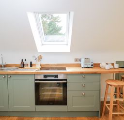 A kitchen with green cabinets wooden countertops a sink a stovetop an oven a toaster cups two wooden stools and a skylight at The Rookery in Fulbrook near Burford