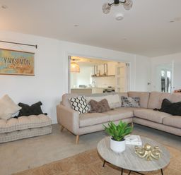 A living room with a beige sectional sofa and cushions a low cushioned bench with pillows a round coffee table with a plant and decoration a view into a kitchen at Ysgol Fawr Big School in Benllech
