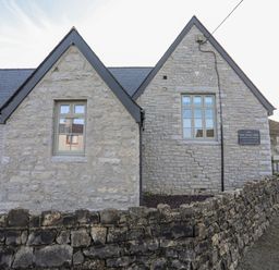 A stone building with pointed roofs and windows behind a stone wall at Ysgol Fawr Big School in Benllech