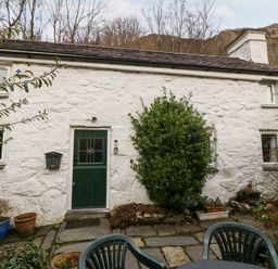 A stone cottage with a green door and windows a bush and potted plants outside with a plastic patio table and chairs at Is Y Graig in Beddgelert