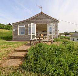 A wooden chalet with open doors and a picnic table outside on a grassy area at Siesta Chalet in Eype Mouth Chalet Park