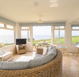 A living room with wicker furniture a TV and large windows overlooking a grassy area and sea at Siesta Chalet in Eype Mouth Chalet Park