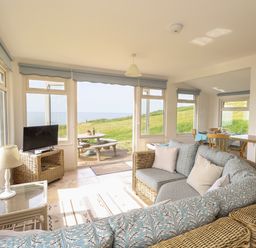 A living room with wicker furniture a television and dining area with outdoor view at Siesta Chalet in Eype Mouth Chalet Park