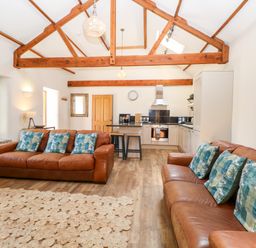 An open-plan living area with brown leather sofas, blue patterned cushions, a kitchen with white cabinets, wooden beams on the ceiling, and a wooden floor at The Barnhouse Felmingham near North Walsham