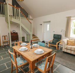 A dining and living area with a wooden table set for four chairs a sofa armchair a staircase and a window at Ystabl Robyn in Llanddarog