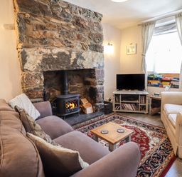 A living room with a stone fireplace a wood stove a beige armchair a brown sofa a wooden coffee table a red patterned rug a tv on a stand and board games by the window at Llygoden Cottage in Beddgelert