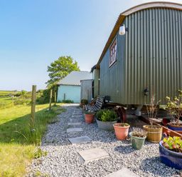 An outdoor space with a corrugated metal hut on wheels a gravel path with potted plants and a bench in a rural area at Puddle Duck Shepherds Hut in Crackington Haven