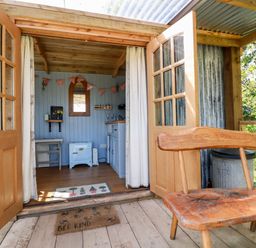 A wooden bench outside open double doors leading to a small room with a light blue stove and kitchen area at Shepherds Retreat Maxworthy near Crackington Haven