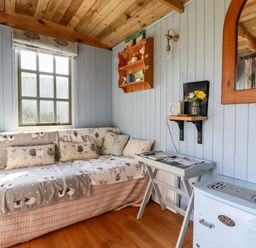 A small room with a wooden ceiling a window a sofa with sheep patterned covers a small table a wall shelf with decorations a mirror and a blue stove at Shepherds Retreat Maxworthy near Crackington Haven