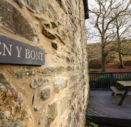 A stone wall with a sign reading Pen y Bont and a wooden deck with a picnic table near trees and hills at Pen y Bont Near Mallwyd