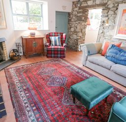 A living room with a red patterned rug a beige sofa with cushions a teal armchair and ottoman a stone wall and a small fireplace at The Old Rectory Coach House in Rathmullan County Donegal
