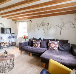 A living room with a dark sofa, patterned cushions, a yellow chair, a coffee table with wine and glasses, wooden beams on the ceiling, and decorative branches on the wall at Heron House in Talley near Llandeilo