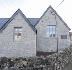 A stone building with two peaked roofs and windows behind a stone wall at Ysgol Fach Little School in Benllech