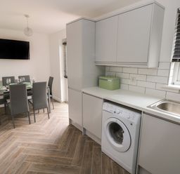 A kitchen with a washing machine under the counter a sink window with blinds and a dining table with six chairs and a wall mounted television at 39 Mariners Quay in Port Talbot