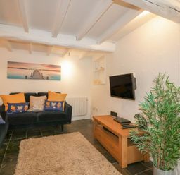 A living room with black sofas yellow and floral cushions a wall-mounted TV and a potted plant at Blacksmiths Cottage in Crantock