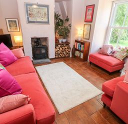 A living room with red sofas pink pillows a white rug a wood stove firewood stacked a bookshelf and a window showing greenery at Pentira in Kuggar