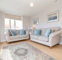 A living room with two beige sofas blue and brown cushions a patterned rug and framed pictures on the wall at 23 Tibby's Way in Southwold
