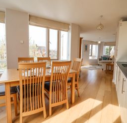 A dining area with a wooden table and six chairs next to large windows and an open living space with sofas at 23 Tibby's Way Southwold
