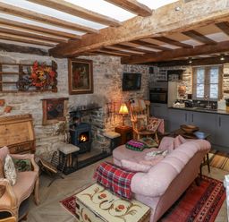A living area with a pink sofa a wood burning stove wooden beams stone walls and a kitchen area at Hoarders Cottage in Hay-On-Wye