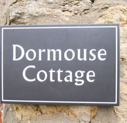 A black sign with white letters reading Dormouse Cottage on a stone wall at Dormouse Cottage Burton Bradstock