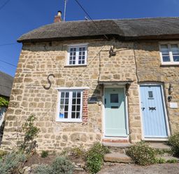A stone cottage with two pastel-colored doors and white framed windows with flowers in front at Dormouse Cottage in Burton Bradstock
