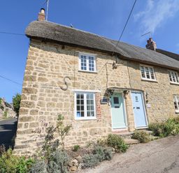 A stone cottage with a thatched roof and two pastel colored doors beside a narrow street at Dormouse Cottage in Burton Bradstock