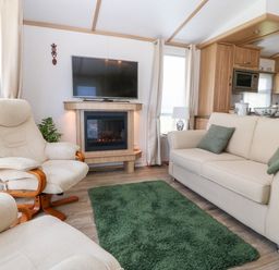 A living room with two beige armchairs a beige sofa green cushions a green rug a wall-mounted TV and a fireplace at Gwel Y Mor in Nasareth near Caernarfon