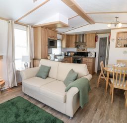 A living area with a cream sofa, green cushions, green rug, kitchen with wooden cabinets, dining table with chairs, and a fireplace at Gwel Y Mor in Nasareth near Caernarfon