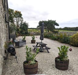 An outdoor patio with gravel ground, wooden picnic table, closed umbrella, two planters with plants, and a stone wall in the background at The Dairy at Brackenthwaite Farm in Arnside