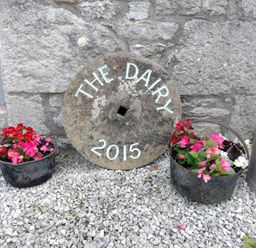 A stone wall with a round stone sign reading The Dairy 2015 between two metal baskets with flowers and a metal milk canister on gravel at The Dairy at Brackenthwaite Farm in Arnside