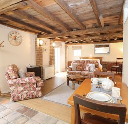 A living area with floral armchair and sofa a wooden dining table set with dishes a bed in the background and a wooden beamed ceiling at Pear Tree Barn in Stalham